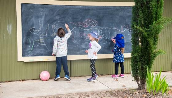 Photo of three children writing on blackboard outside at kindy